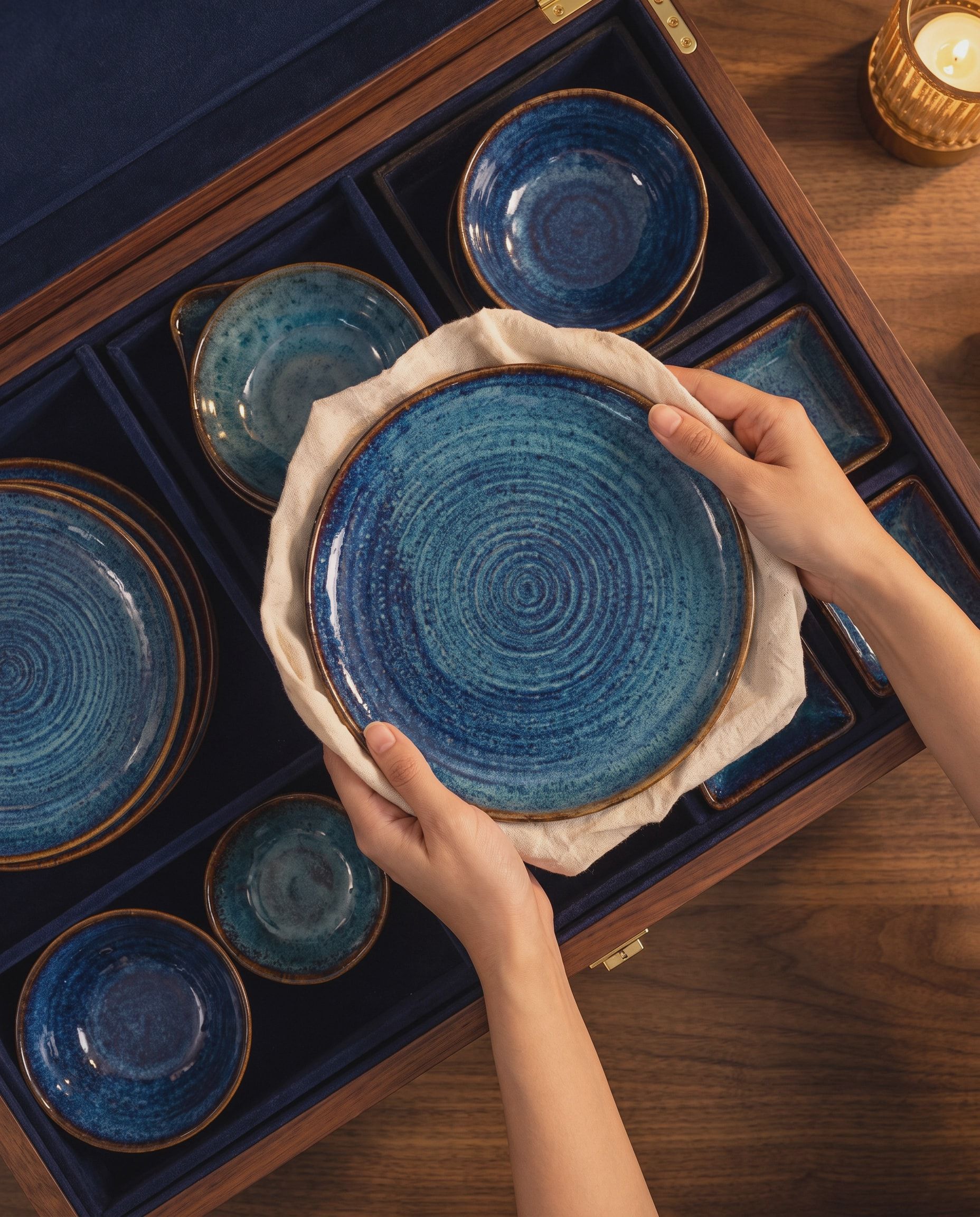 A blue-glazed NEELĀ plate in a linen cloth, held in two hands over the open chest, bowls, plates and dishes in their navy velvet places beneath